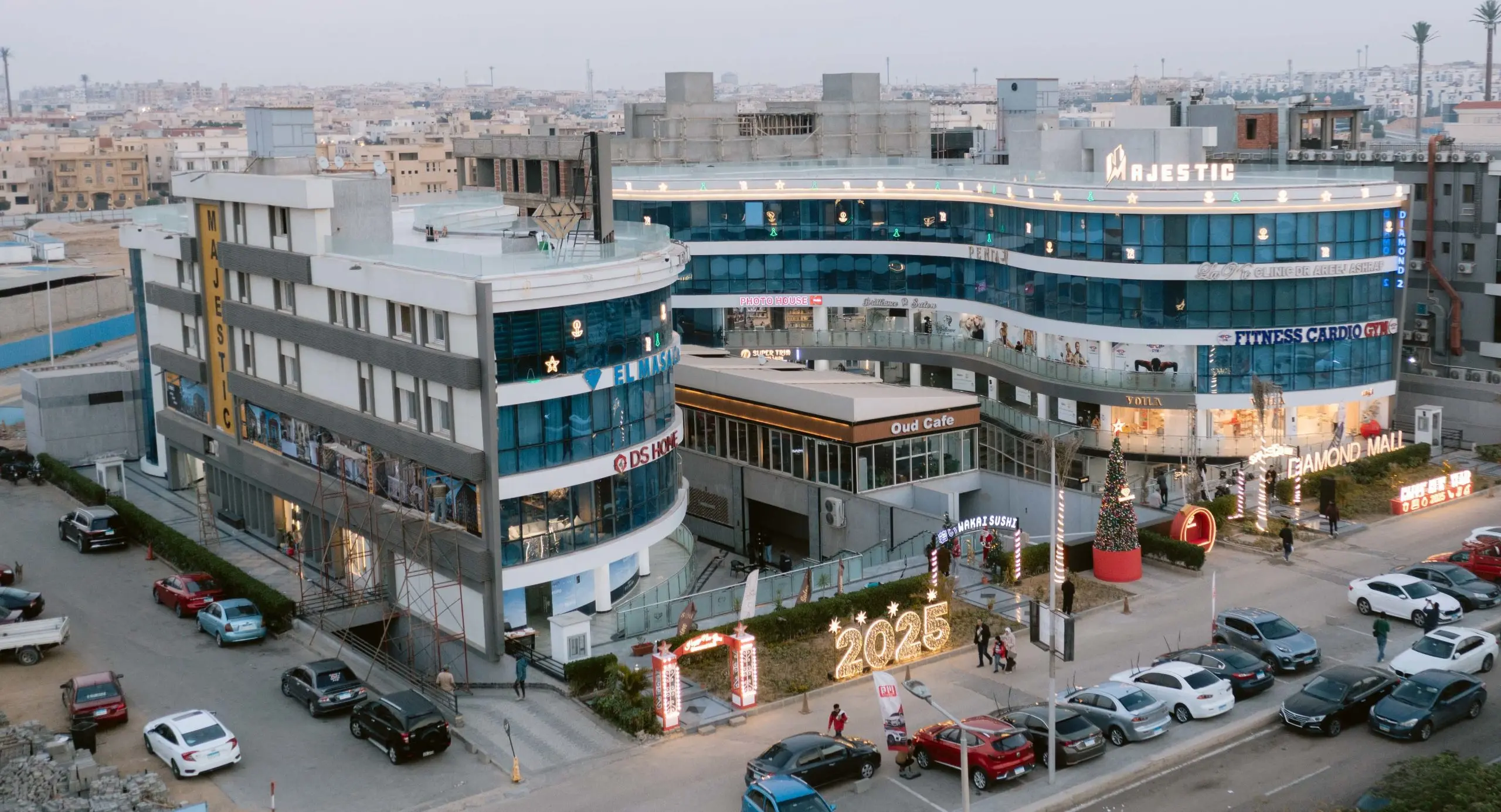 Aerial view of DIAMOND MALL at dusk with festive lighting and a Christmas tree in the foreground, and a full parking lot - دايموند مول.